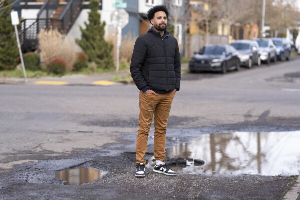 Timothy Taylor poses for a photo by pot holes outside his home on Thursday, Feb. 6, 2025, in Portland, Ore. Taylor said a pot hole broke part of his car's suspension and cost him $1,000 to fix. (AP Photo/Jenny Kane)